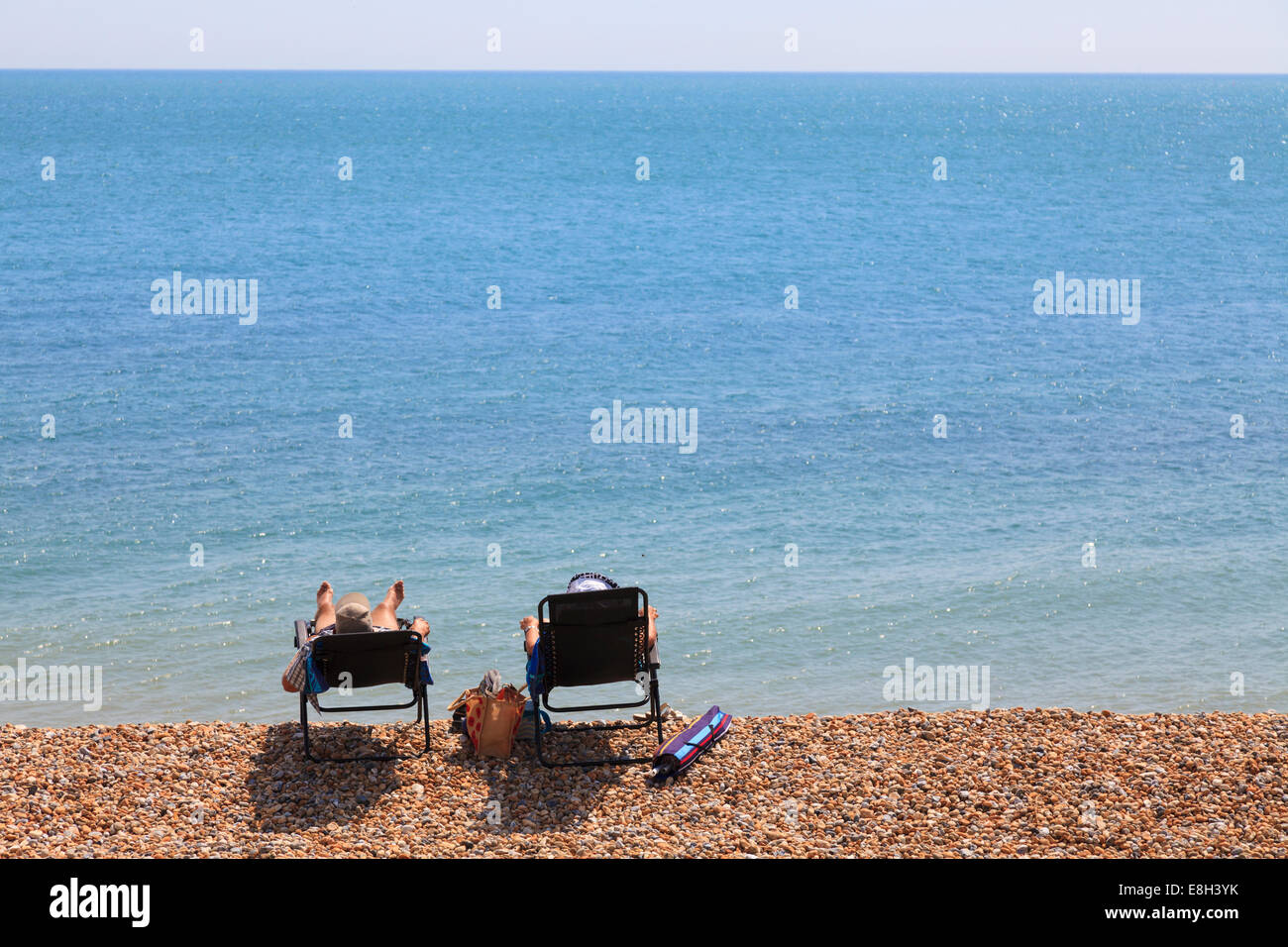 Two people in sun loungers on shingle beach by sea from behind. Stock Photo