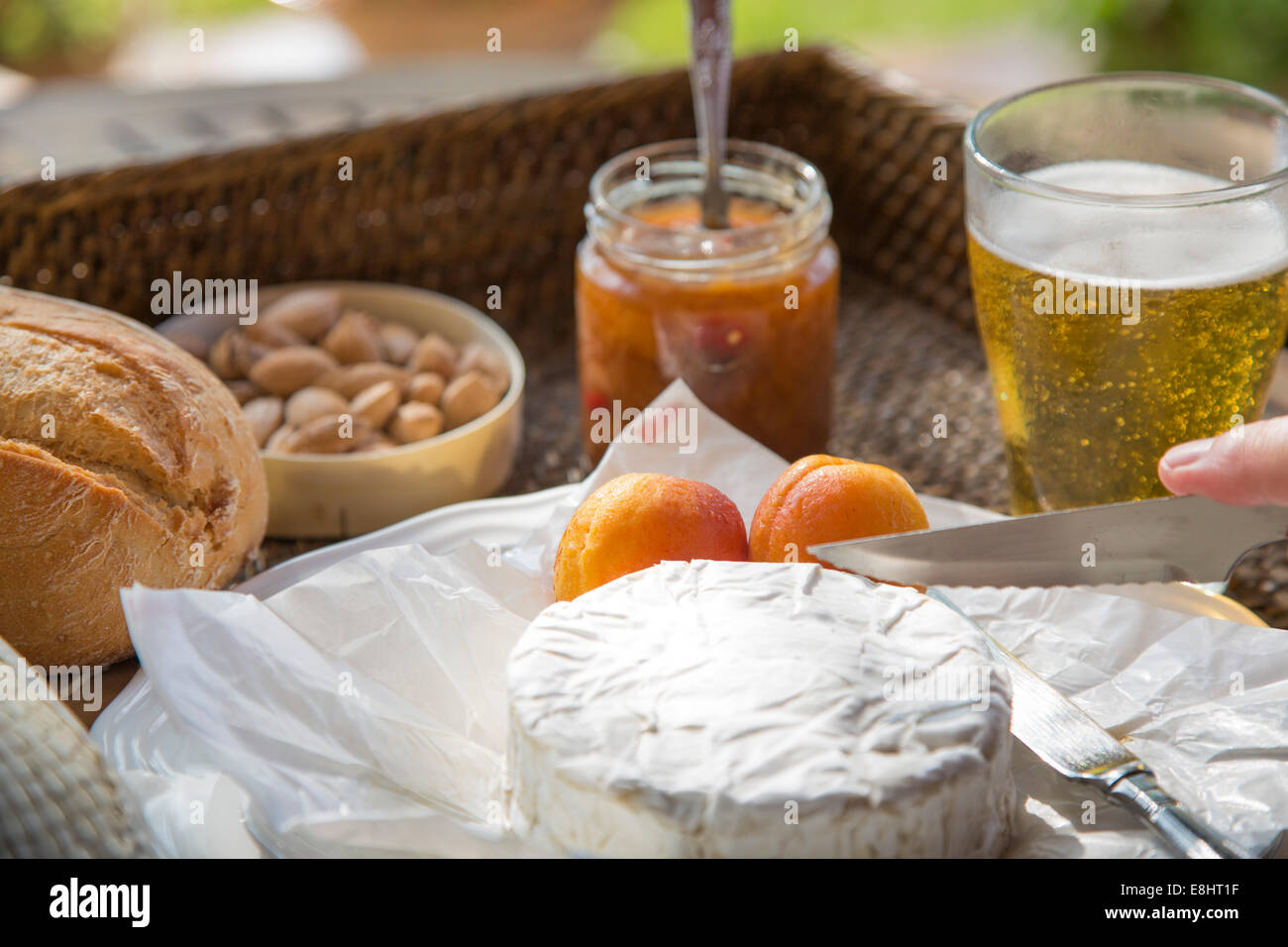 camembert cheese being cut, with apricots, apricot chutney, almonds and beer, set on woven tray Stock Photo