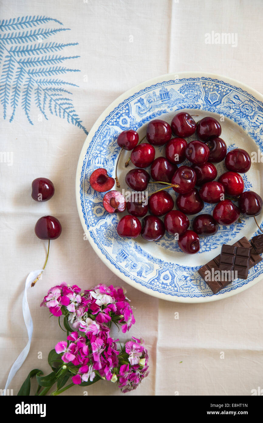 whole cherries and chocolate on blue ceramic plate, with leaf motif table linen and flowers Stock Photo
