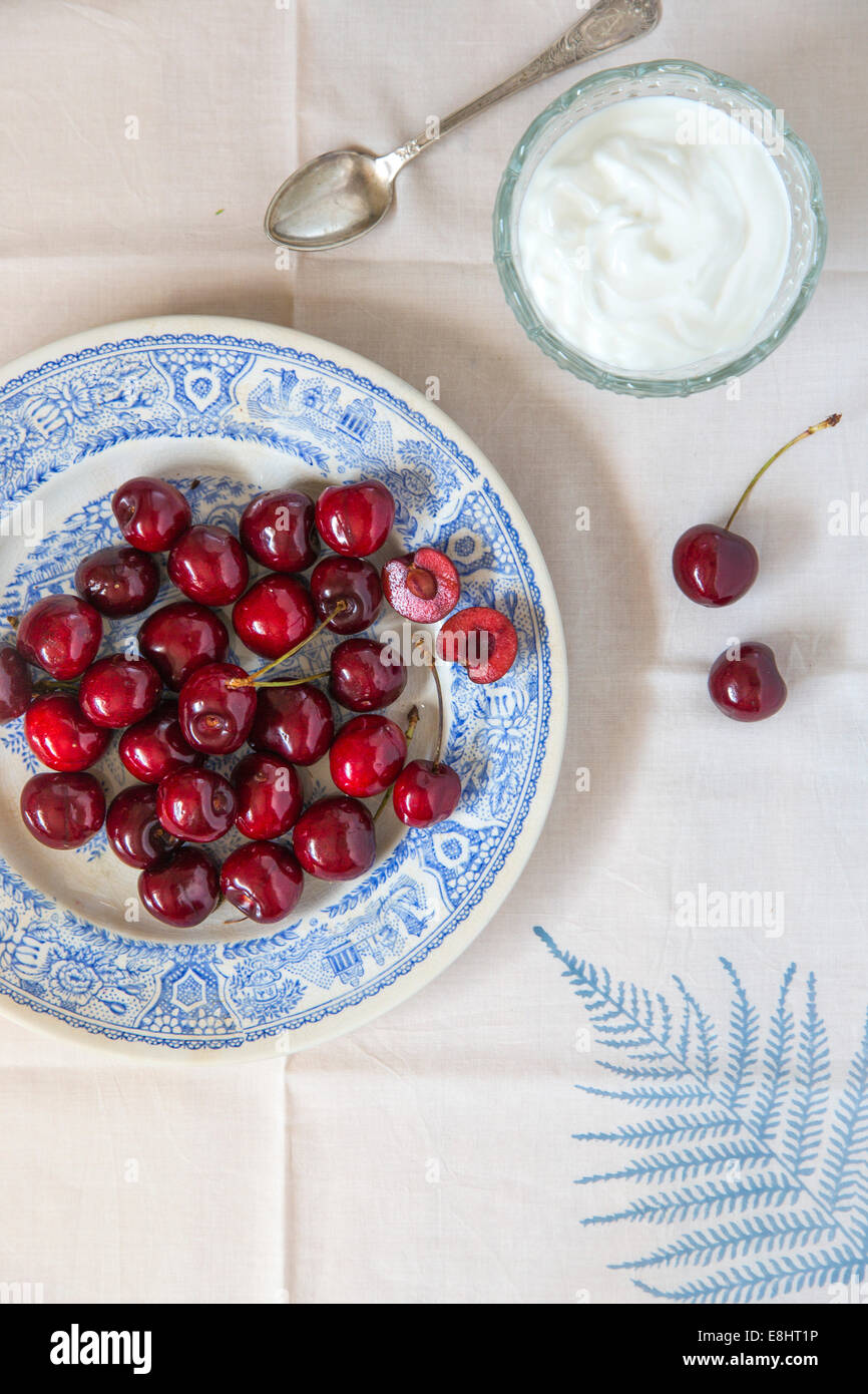 whole cherries on blue ceramic plate, on leaf linen and cream or yogurt in glass dish with vintage spoon Stock Photo