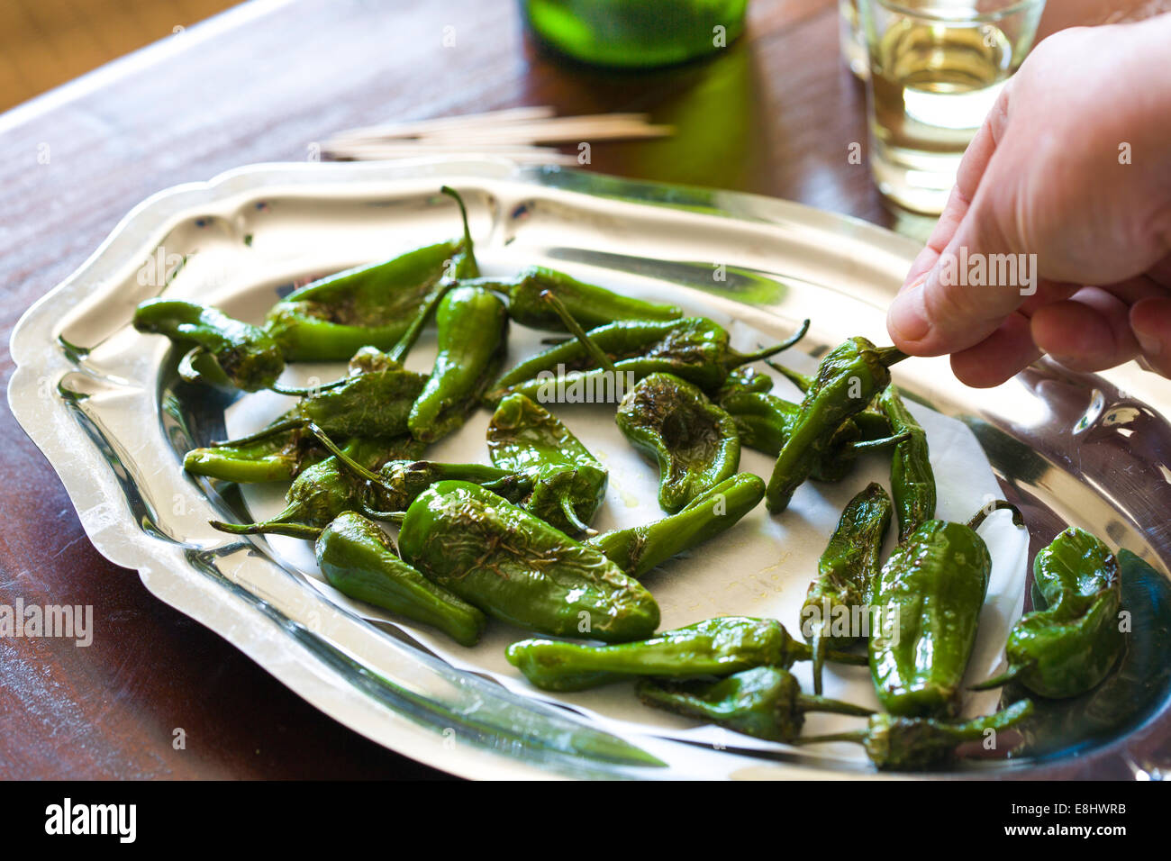 padron peppers as a typical Spanish tapas style snack on wood table , with sherry glasses and bottle in background and hand hold Stock Photo