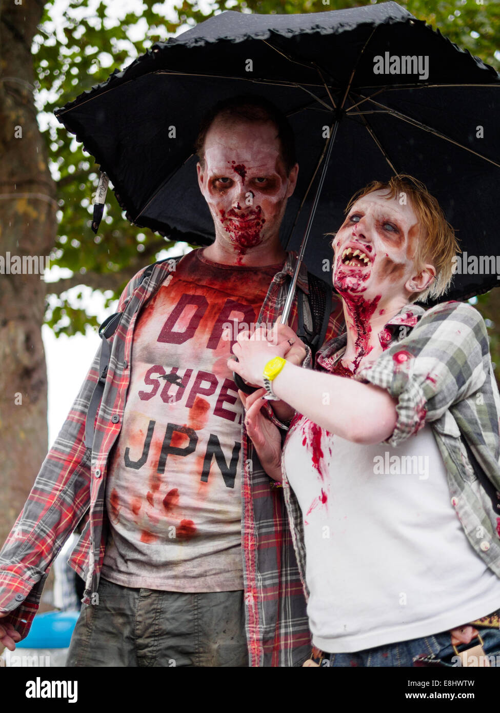 Zombie couple walking under trees in light rain at Southbank, London, holding an umbrella. Stock Photo