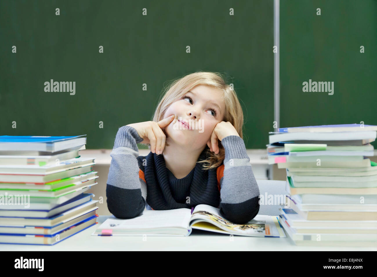 Schoolgirl, 9 years, looking pensive, sitting between two piles of books in front of a blackboard Stock Photo