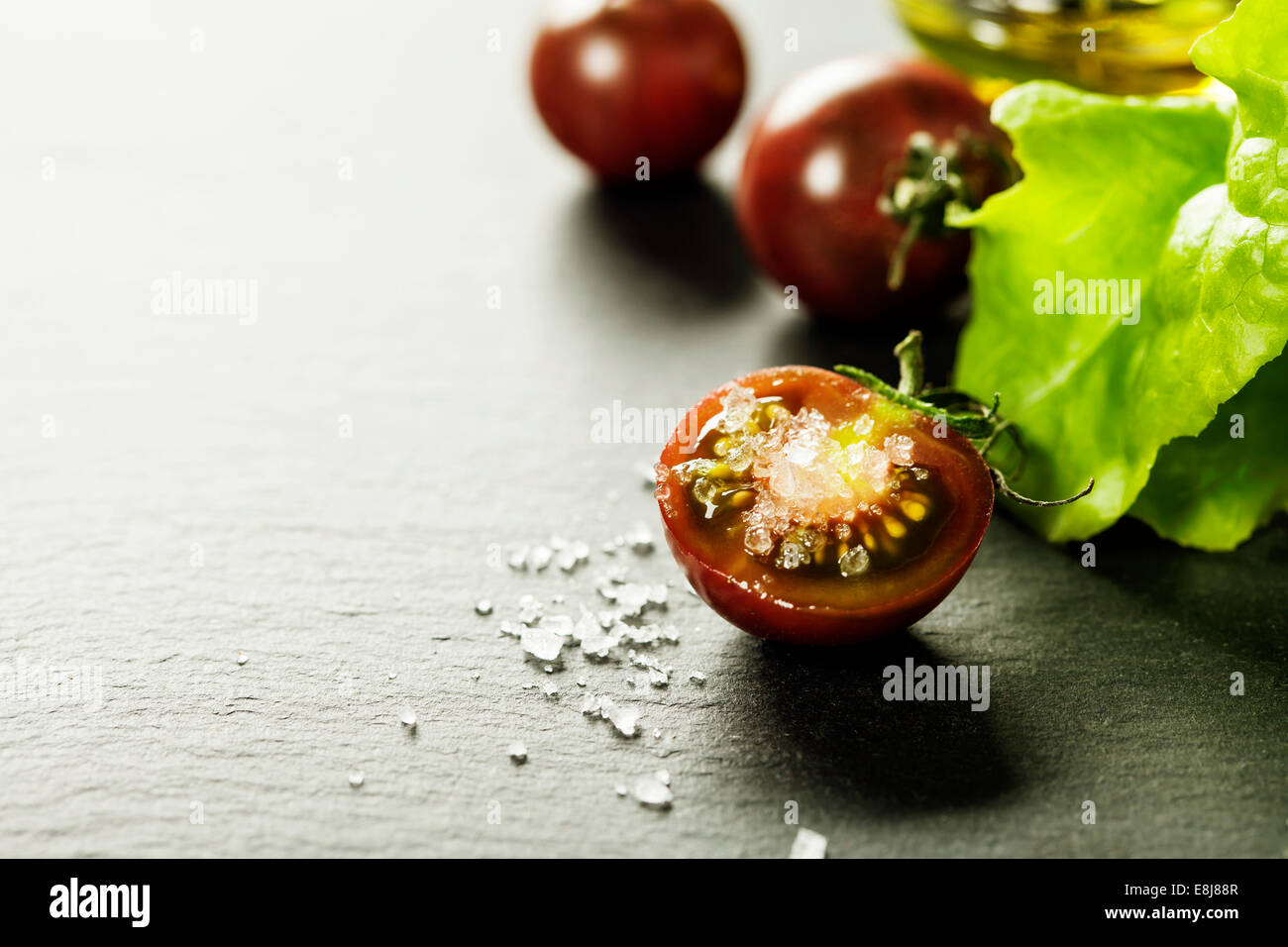 Fresh grape tomatoes with salad leaves and salt for use as cooking ingredients with halved tomato in foreground with copyspace Stock Photo