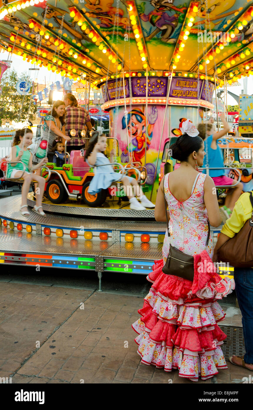 Spanish woman in traditional festive dress waiting next to chlidrens Carousel, merry-go-round at annual fair. Fuengirola, Spain. Stock Photo