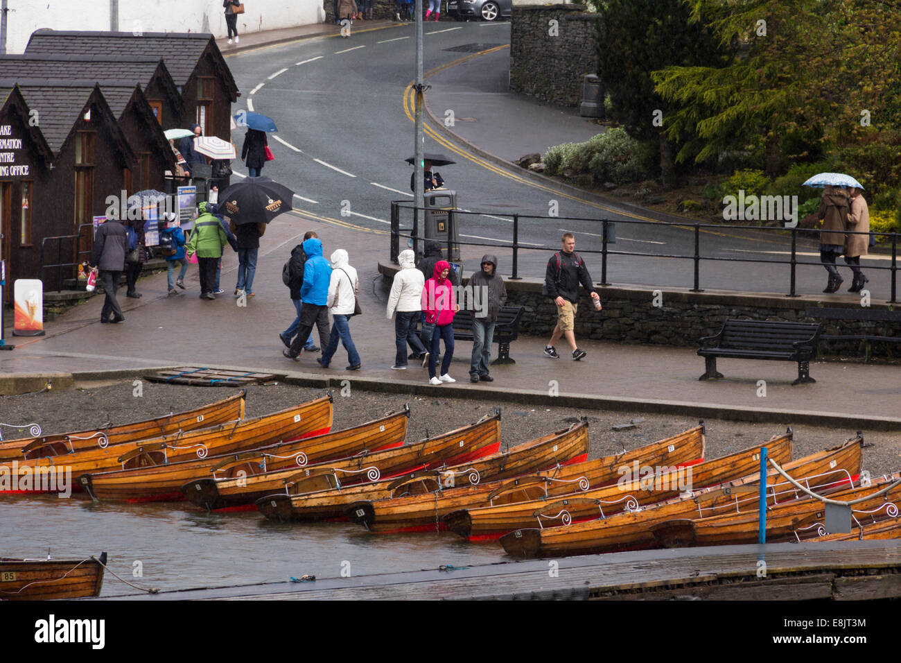 Beached rowing boats and small numbers of  rainwear-clad passers-by near Bowness pier, Cumbria on a very dull and wet day. Stock Photo