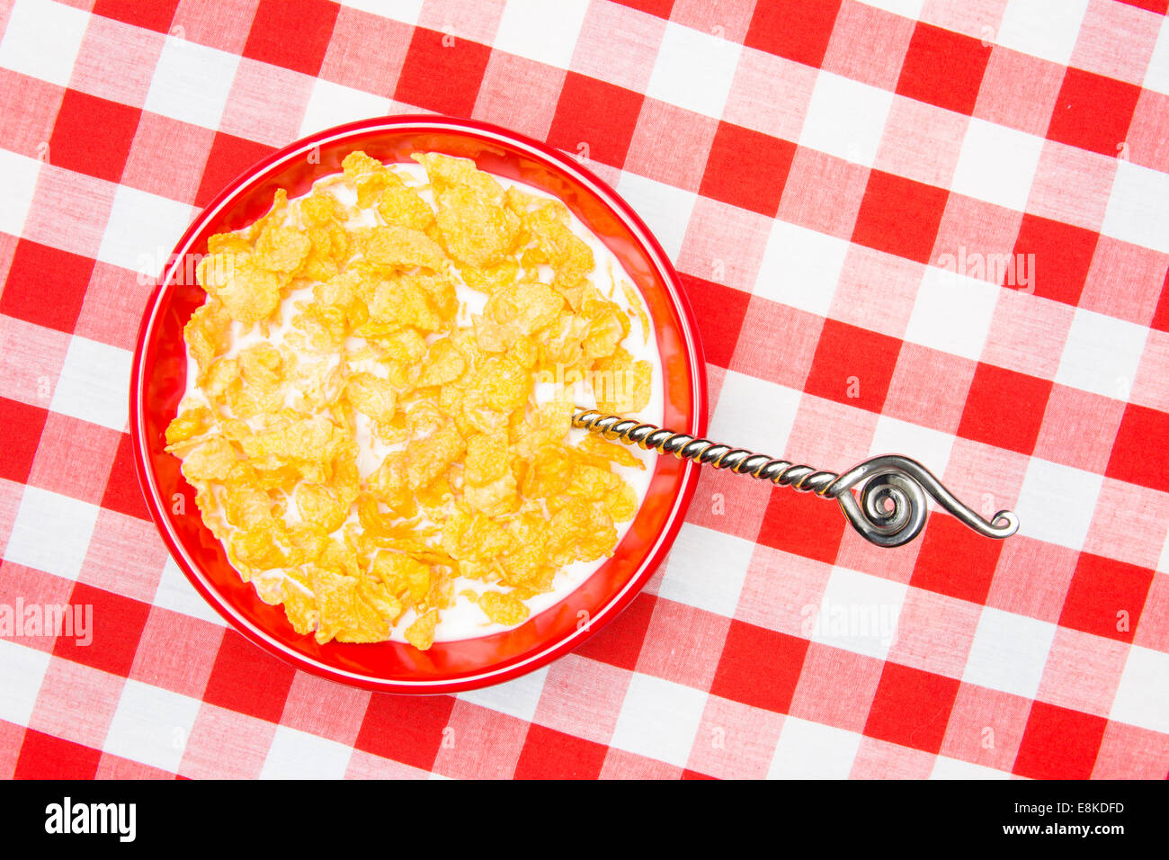 A red bowl of cornflakes and milk on a classic, red, checkered tablecloth ready as a breakfast meal. Stock Photo
