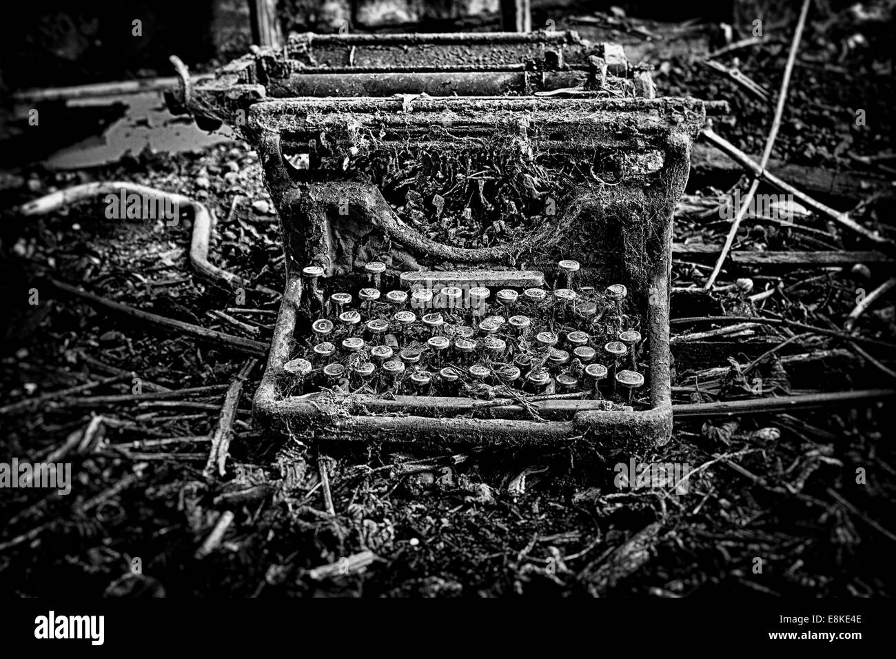 An abandoned and broken, antique, vintage typewriter, left outdoors with weeds and grass growing over it. In black and white. Stock Photo