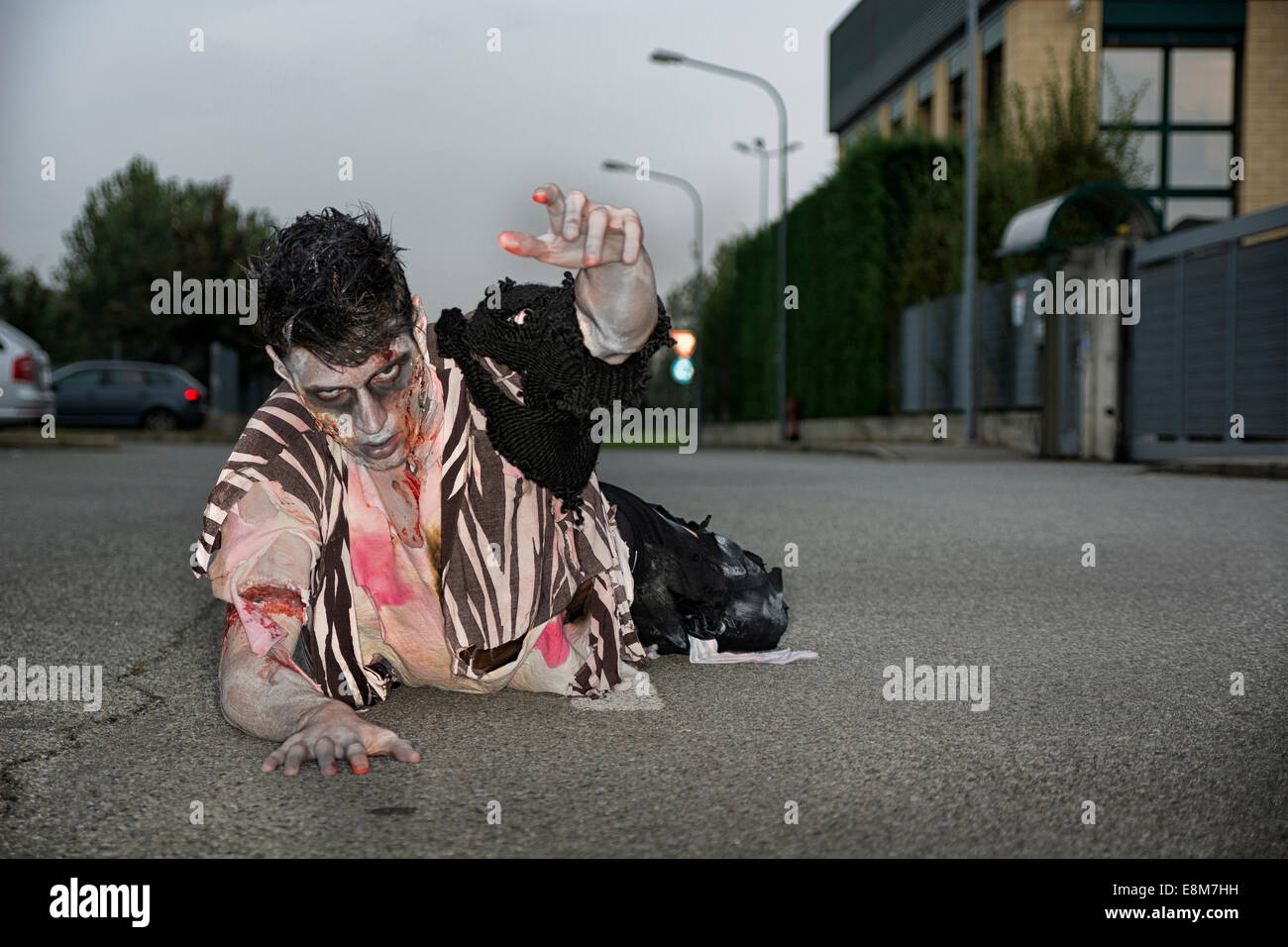 Male zombie crawling on his knees, on empty city street, looking at camera reaching out one hand. Halloween theme Stock Photo