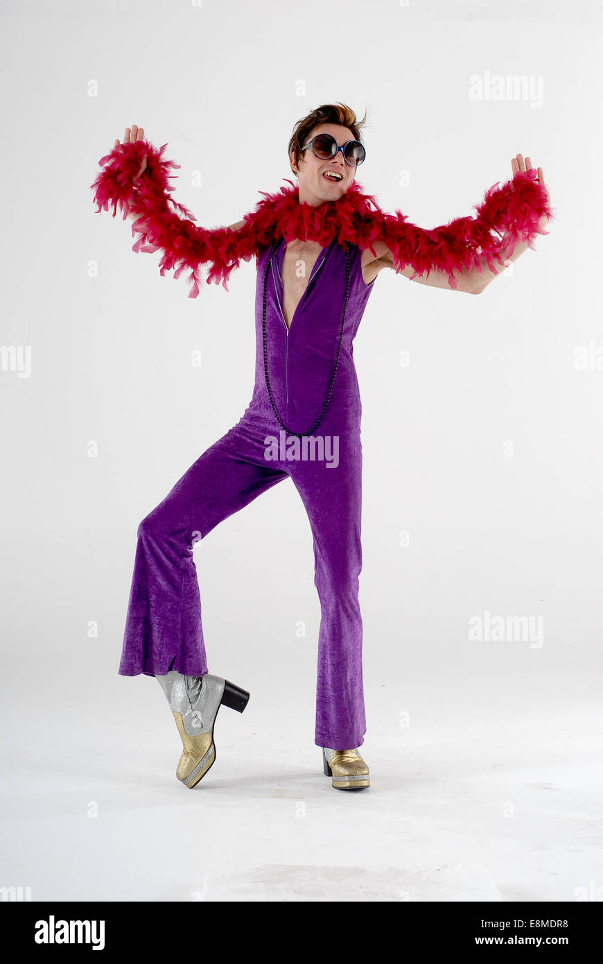 man in fancy dress comedy costume in 1970s flamboyant camp disco outfit  with purple catsuit, platform shoes & feather bower Stock Photo - Alamy