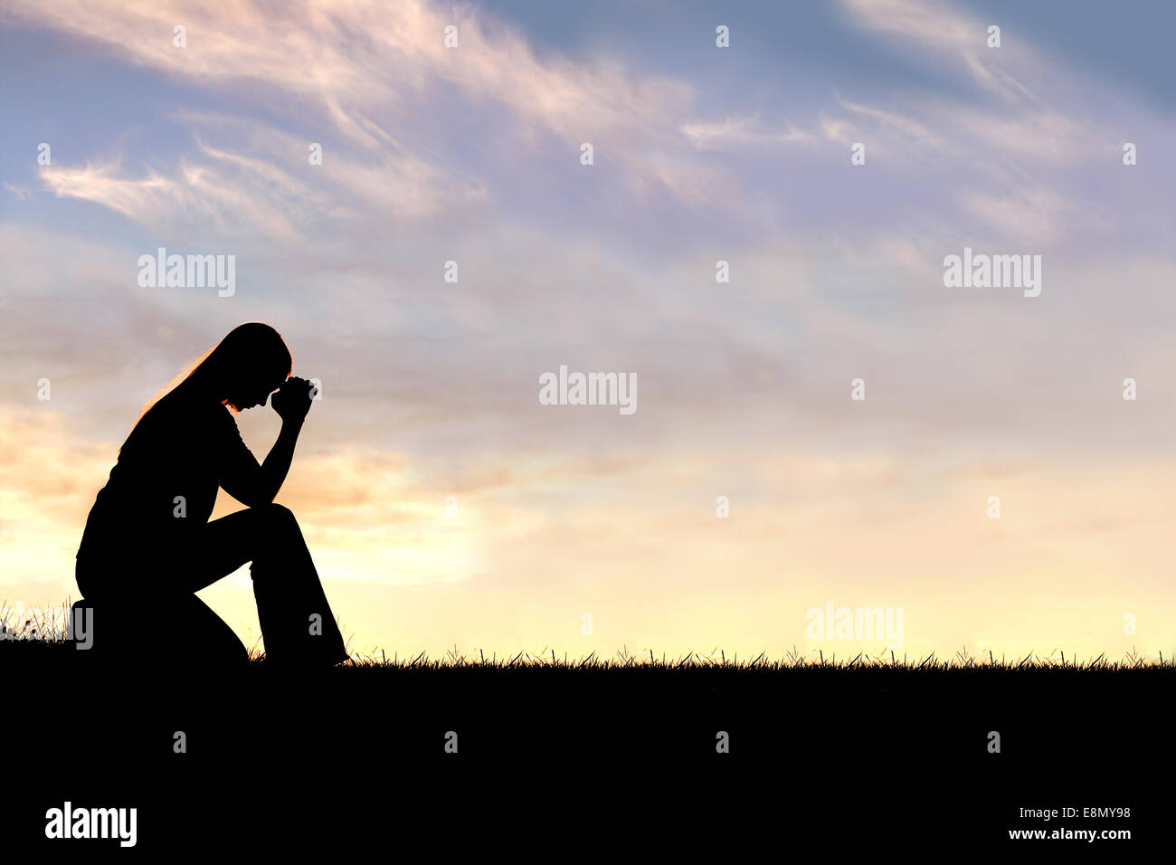 A silhouette of a young Christian woman is bowing her head in prayer, and desperation outside during sunset. Stock Photo