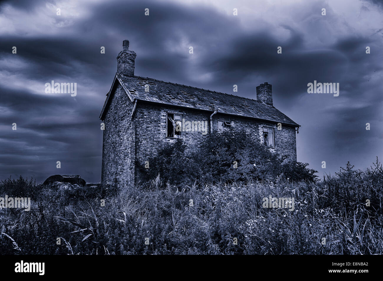 Spooky, haunted, derelict house with broken windows under a stormy sky. Black and white with a blue tint effect. Stock Photo