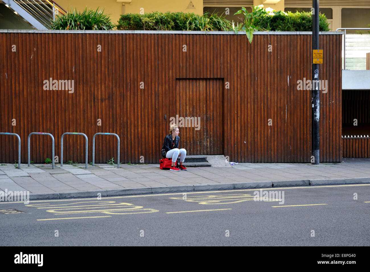 Young woman sitting on red bag by wall next to roadside bus stop Stock Photo