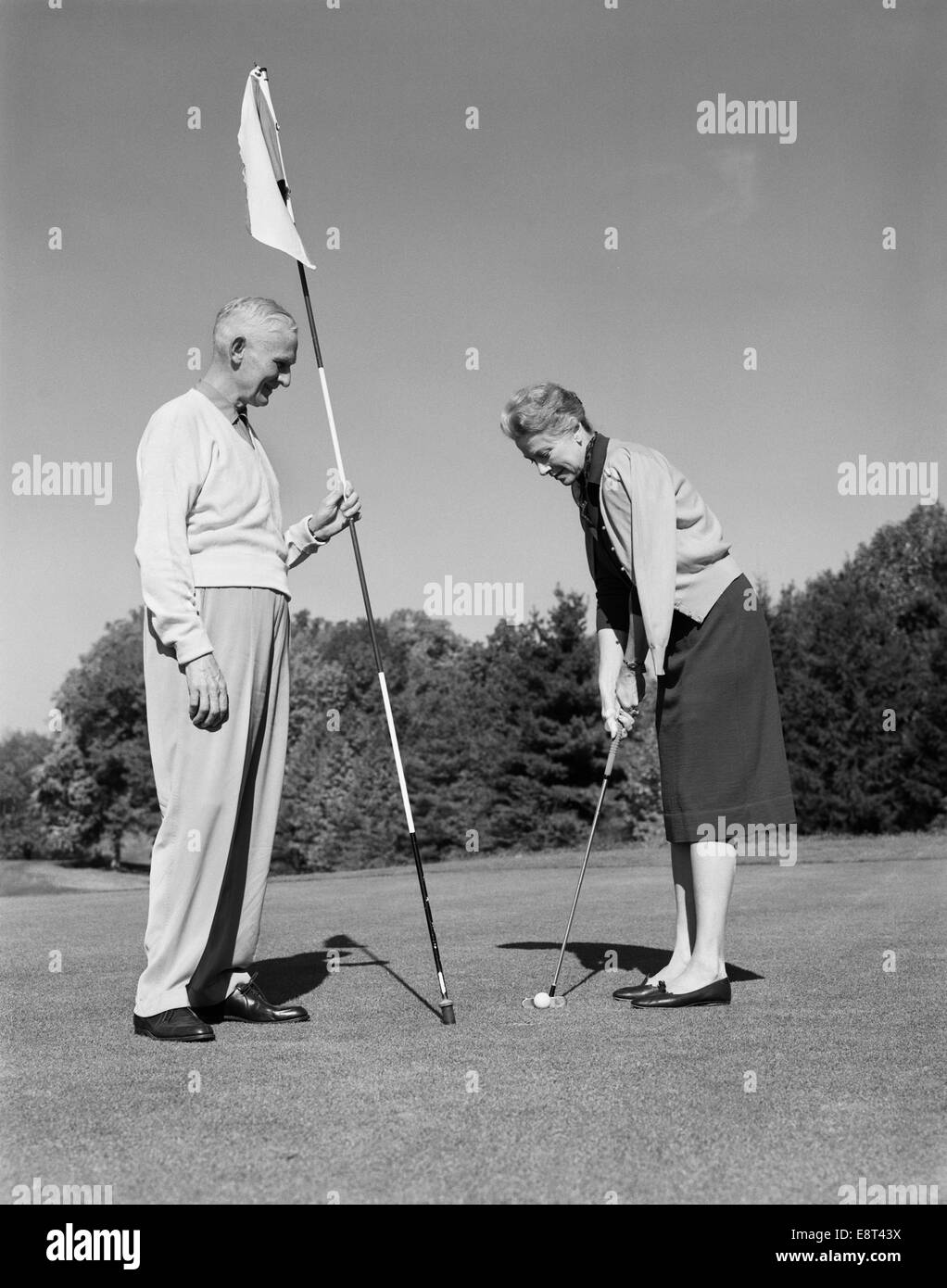 1960s ELDERLY WOMAN GETTING READY TO PUTT WHILE HUSBAND LOOKS ON HOLDING FLAG Stock Photo