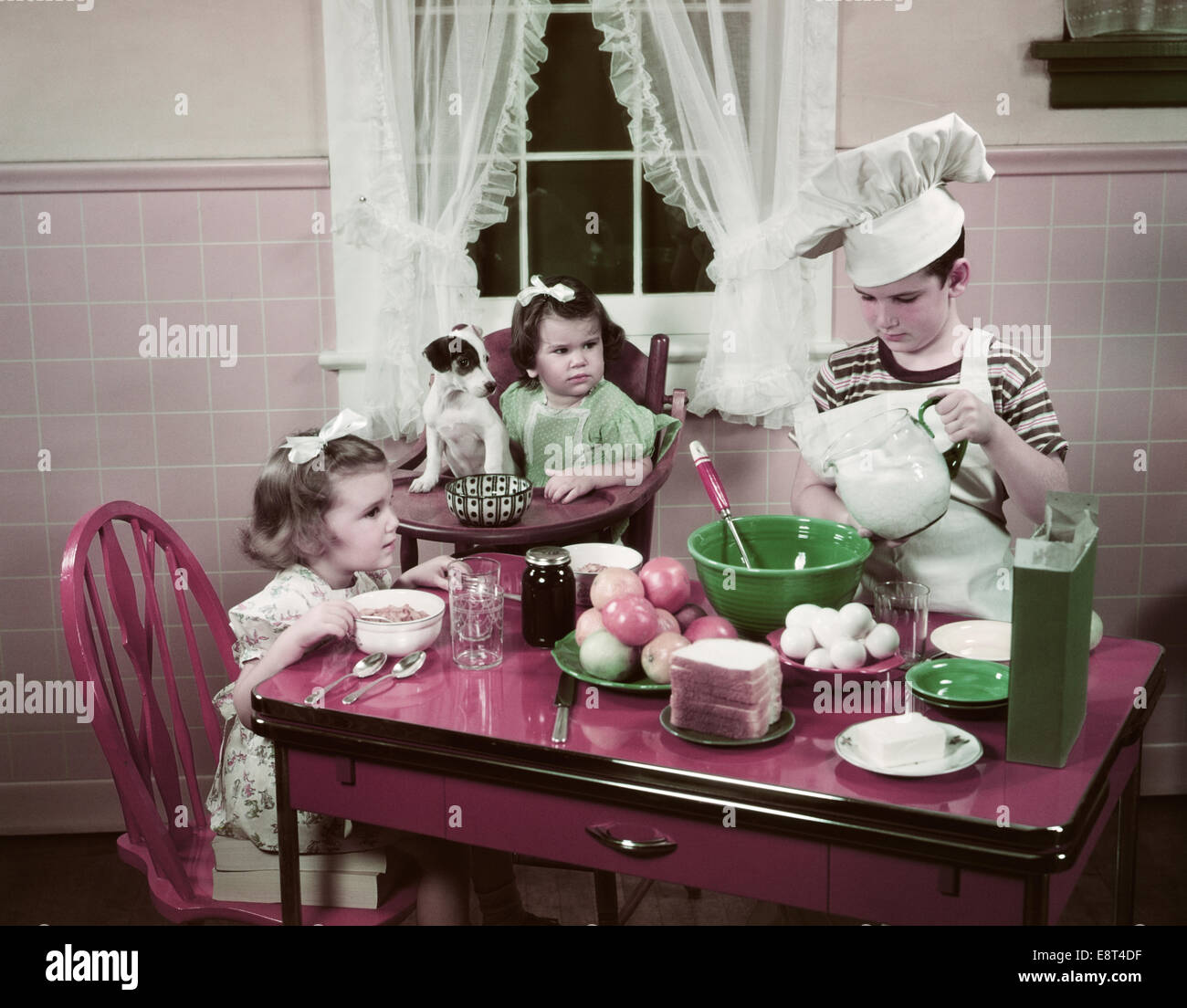 1940s 1950s BOY TWO GIRLS AT KITCHEN TABLE BOY IN TOQUE COOKING ONE GIRL EATING TODDLER AND PUPPY IN HIGH CHAIR Stock Photo