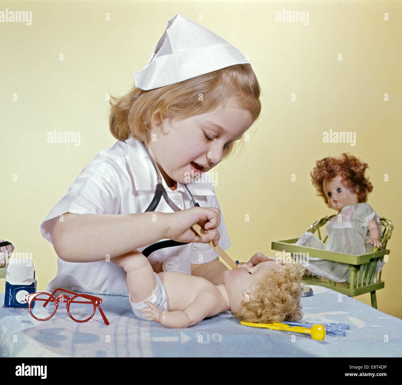 1960s LITTLE GIRL DRESSED UP AS A NURSE TAKING CARE OF HER DOLL Stock Photo