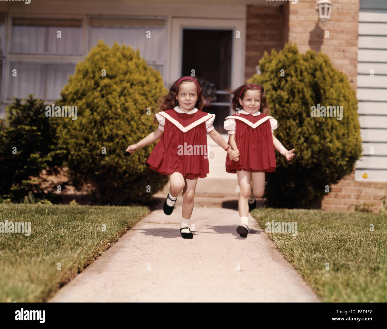 1960s TWIN GIRLS WEARING IDENTICAL RED DRESSES HOLDING HANDS RUNNING DOWN SIDEWALK OF SUBURBAN HOUSE LOOKING AT CAMERA Stock Photo