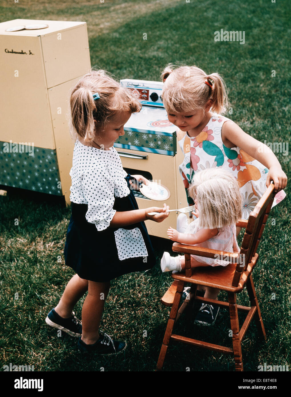 1960s TWO LITTLE GIRLS PLAYING TOGETHER FEEDING DOLL TOY STOVE REFRIGERATOR IN BACKYARD Stock Photo