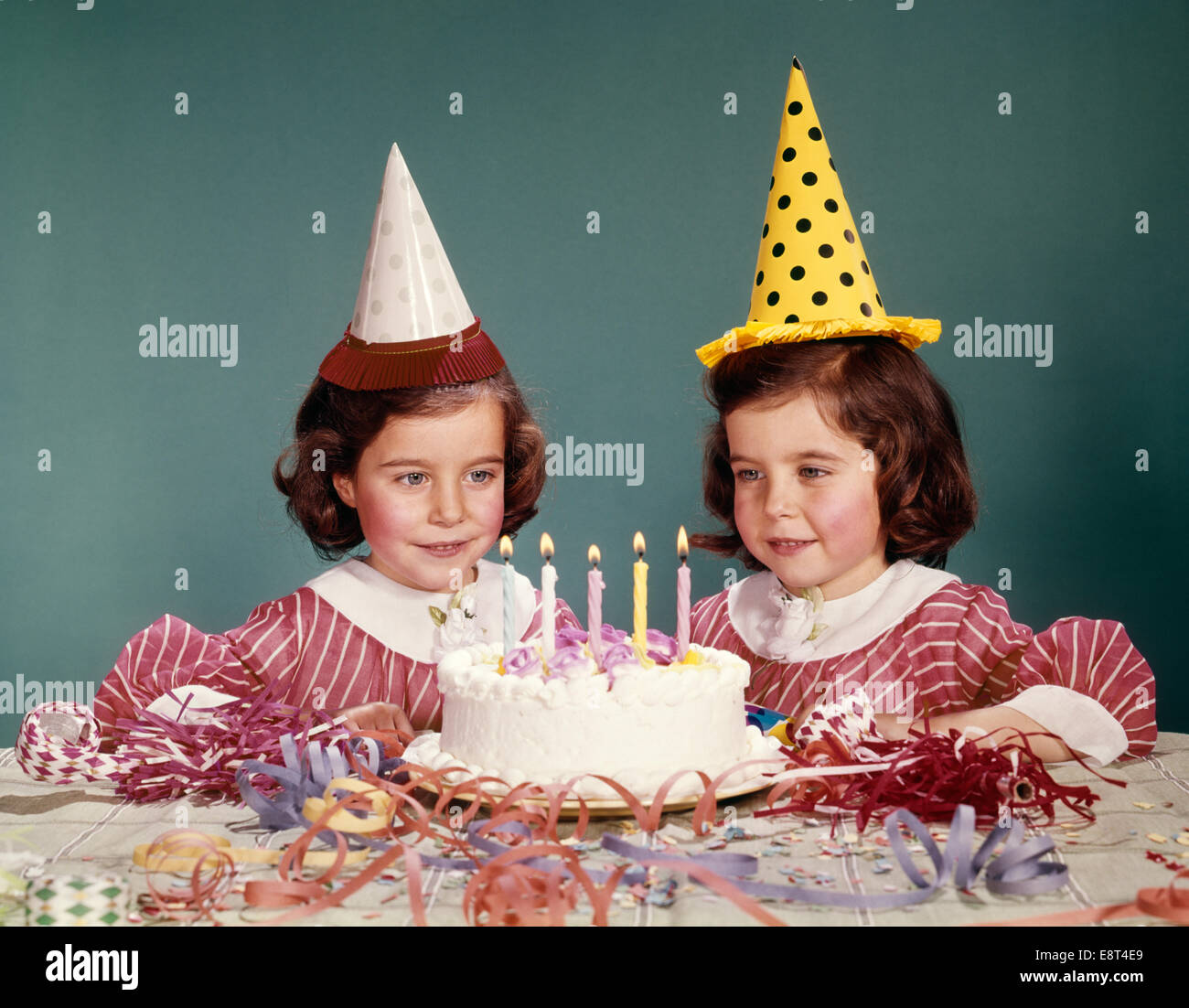 1960s TWIN GIRLS WEARING PARTY HATS AND BIRTHDAY CAKE WITH FIVE CANDLES