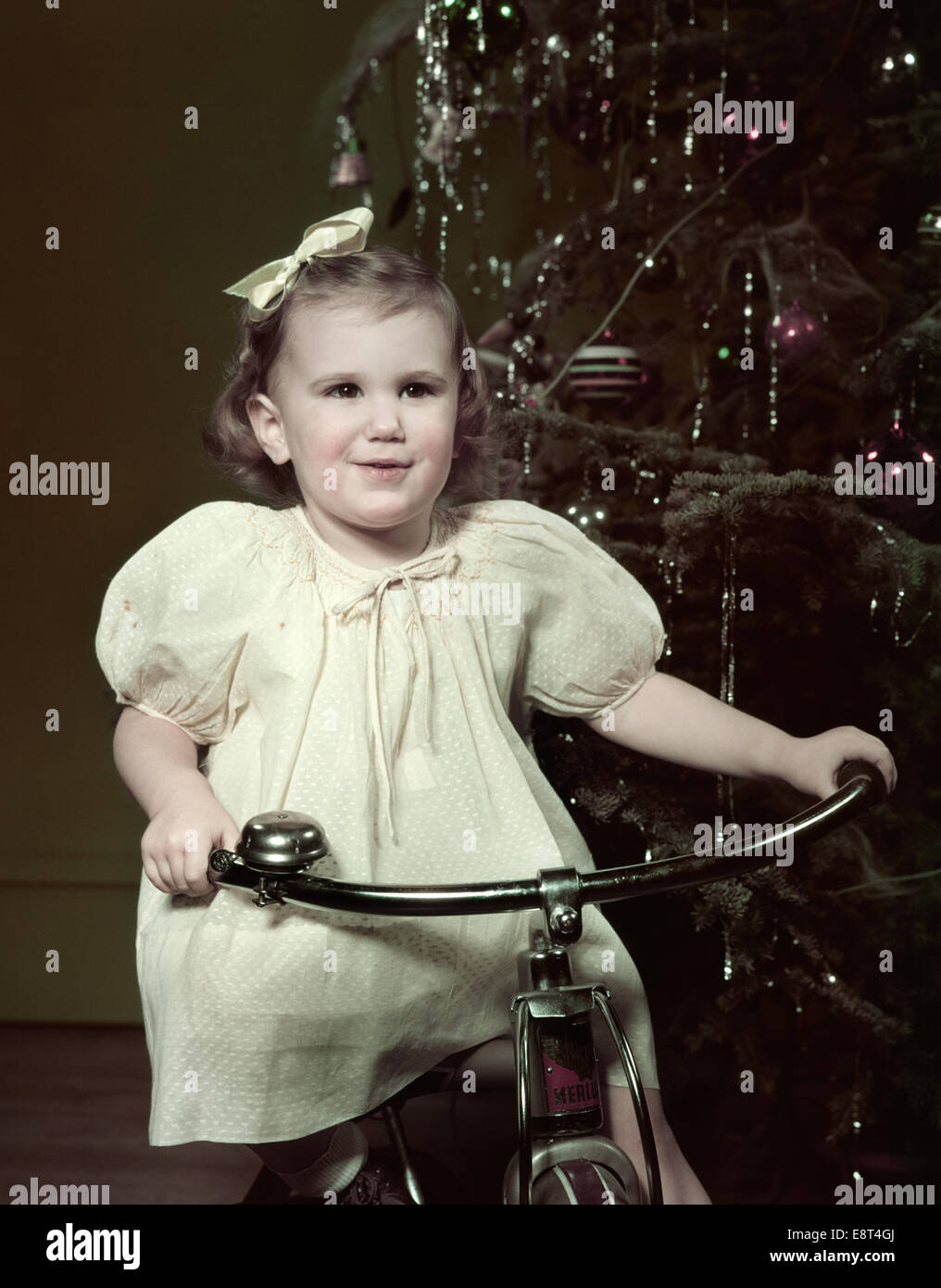 1950s SMILING GIRL RIDING TRICYCLE BY CHRISTMAS TREE Stock Photo