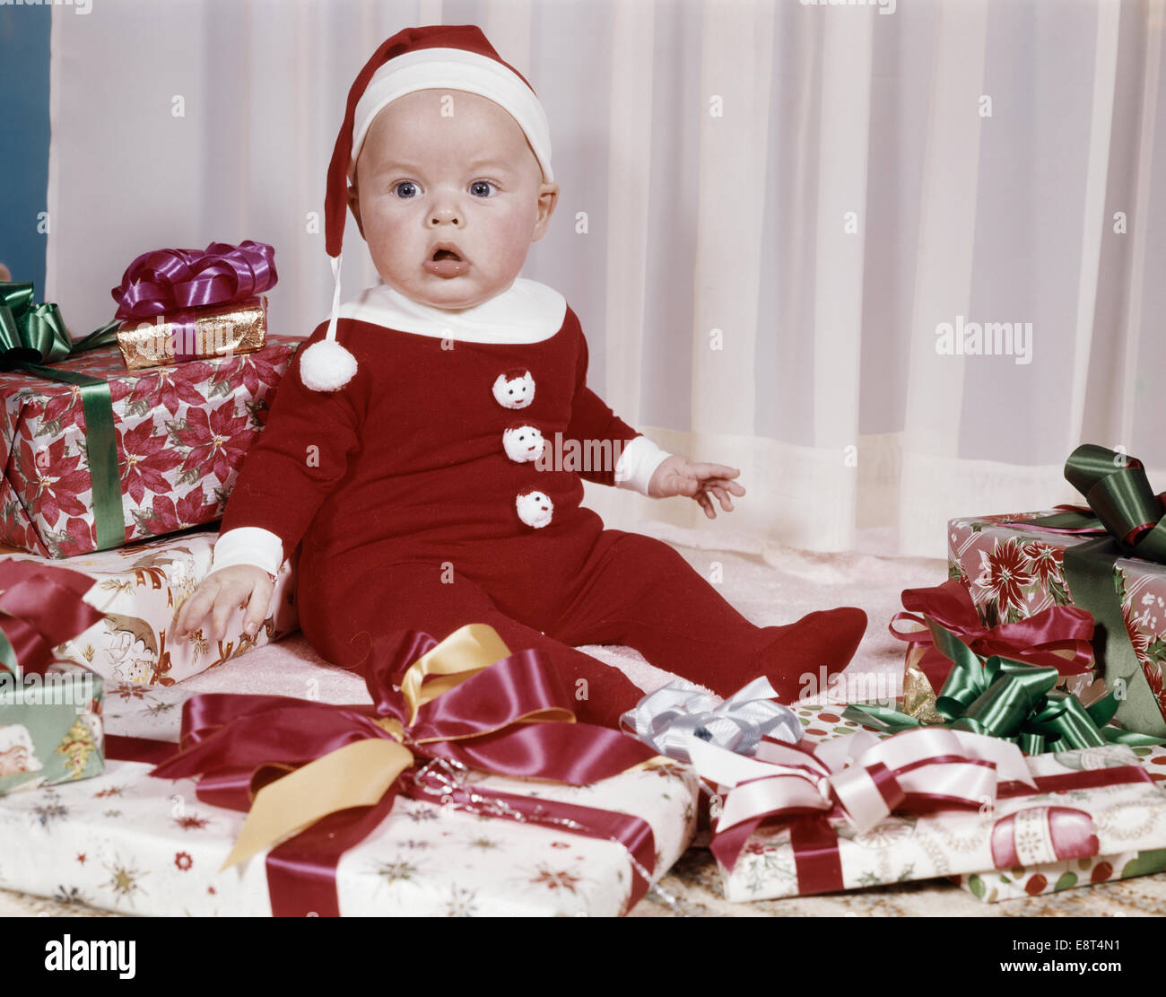 1960s AMAZED BABY IN SANTA SUIT SITTING AMONG WRAPPED PRESENTS Stock Photo