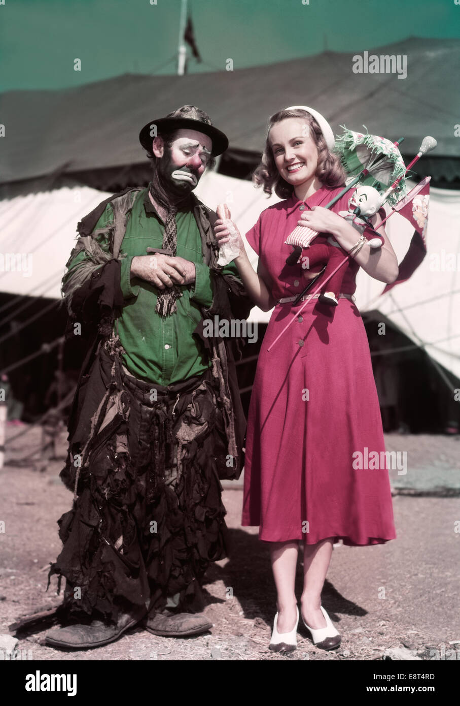 1940s 1950s SMILING BLOND WOMAN AT CIRCUS HOLDING HOT DOG AND PRIZES STANDING NEXT TO HUNGRY SAD MAN HOBO CLOWN EMMETT KELLY Stock Photo