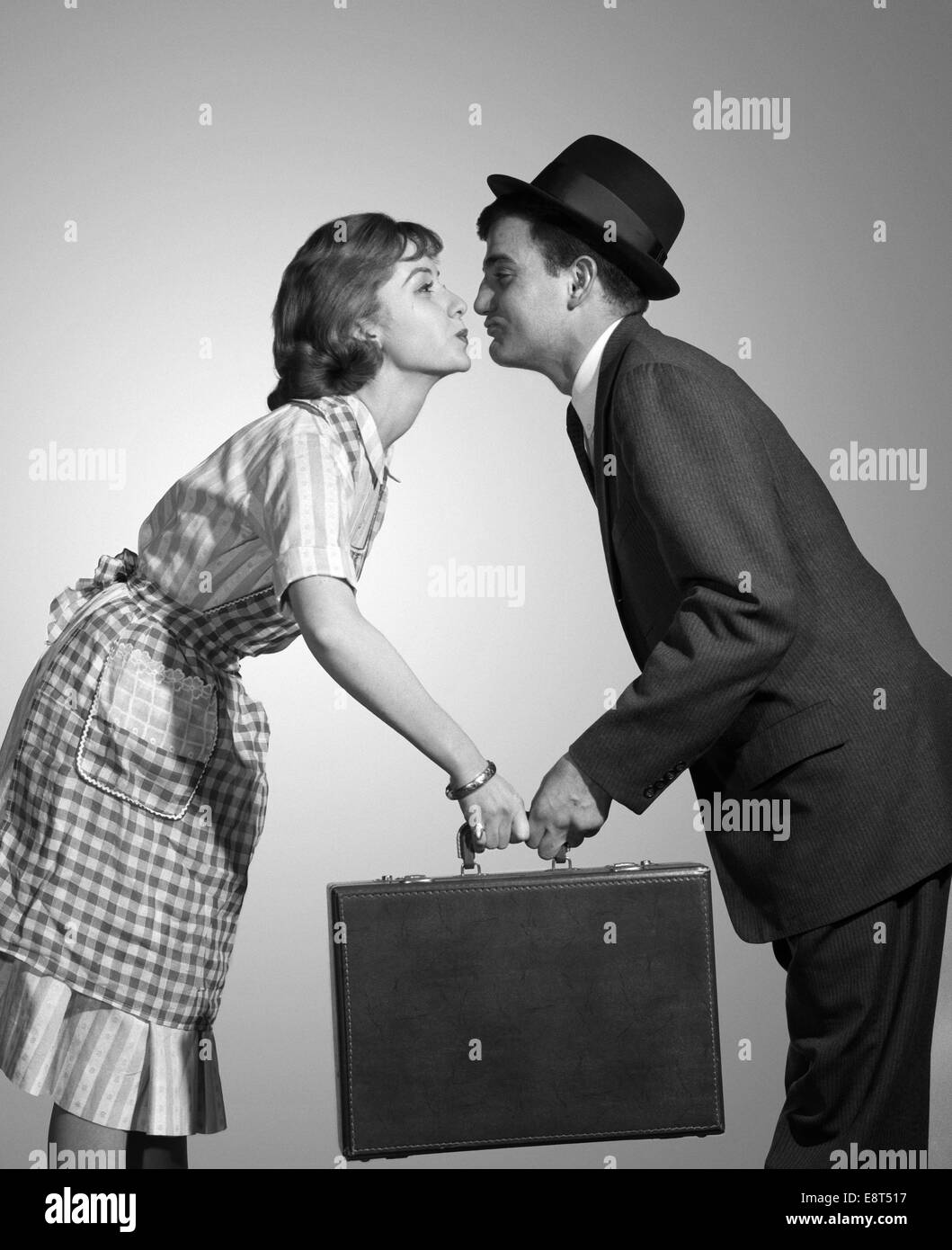 1950s 1960s HOMEMAKER WIFE IN CHECKED APRON KISSING BUSINESSMAN HUSBAND IN SUIT HAT AND TIE AS SHE HANDS HIM A BRIEFCASE Stock Photo