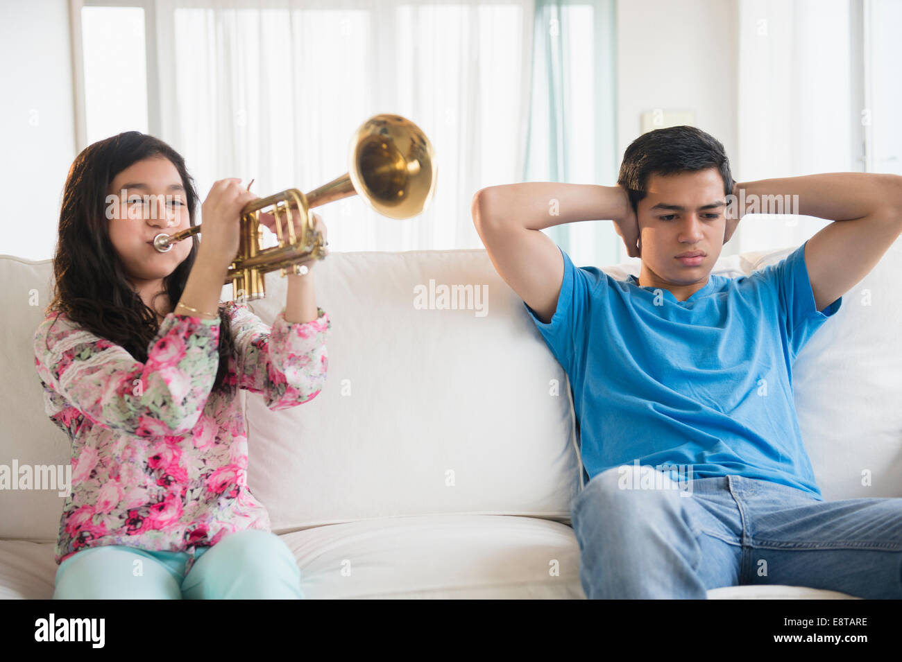 Hispanic brother covering his ears as sister practices trumpet in living room Stock Photo