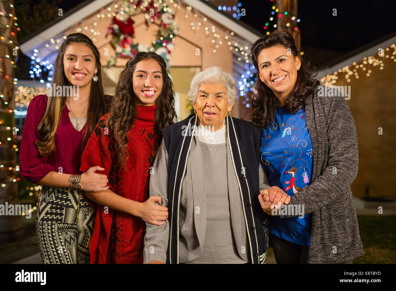Three generations of Hispanic women smiling outside house decorated with string lights Stock Photo