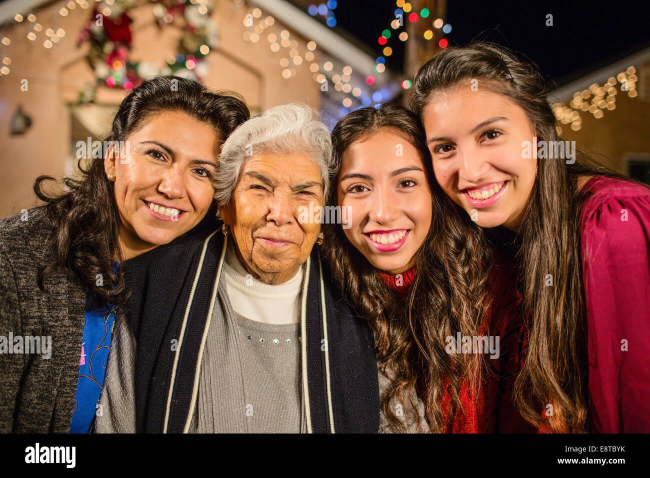 Three generations of Hispanic women standing outside house decorated with string lights Stock Photo