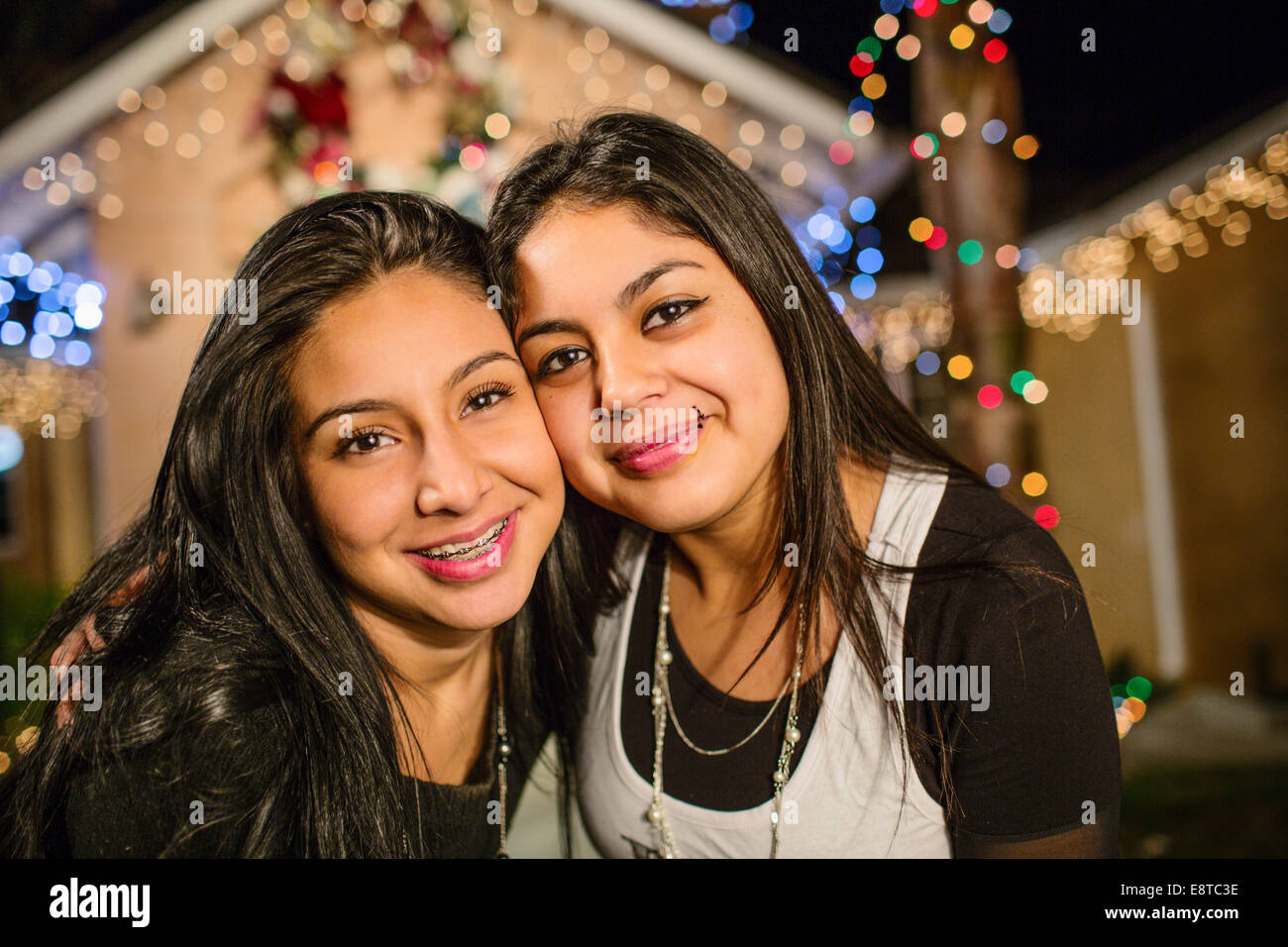 Hispanic sisters smiling outside house decorated with string lights Stock Photo