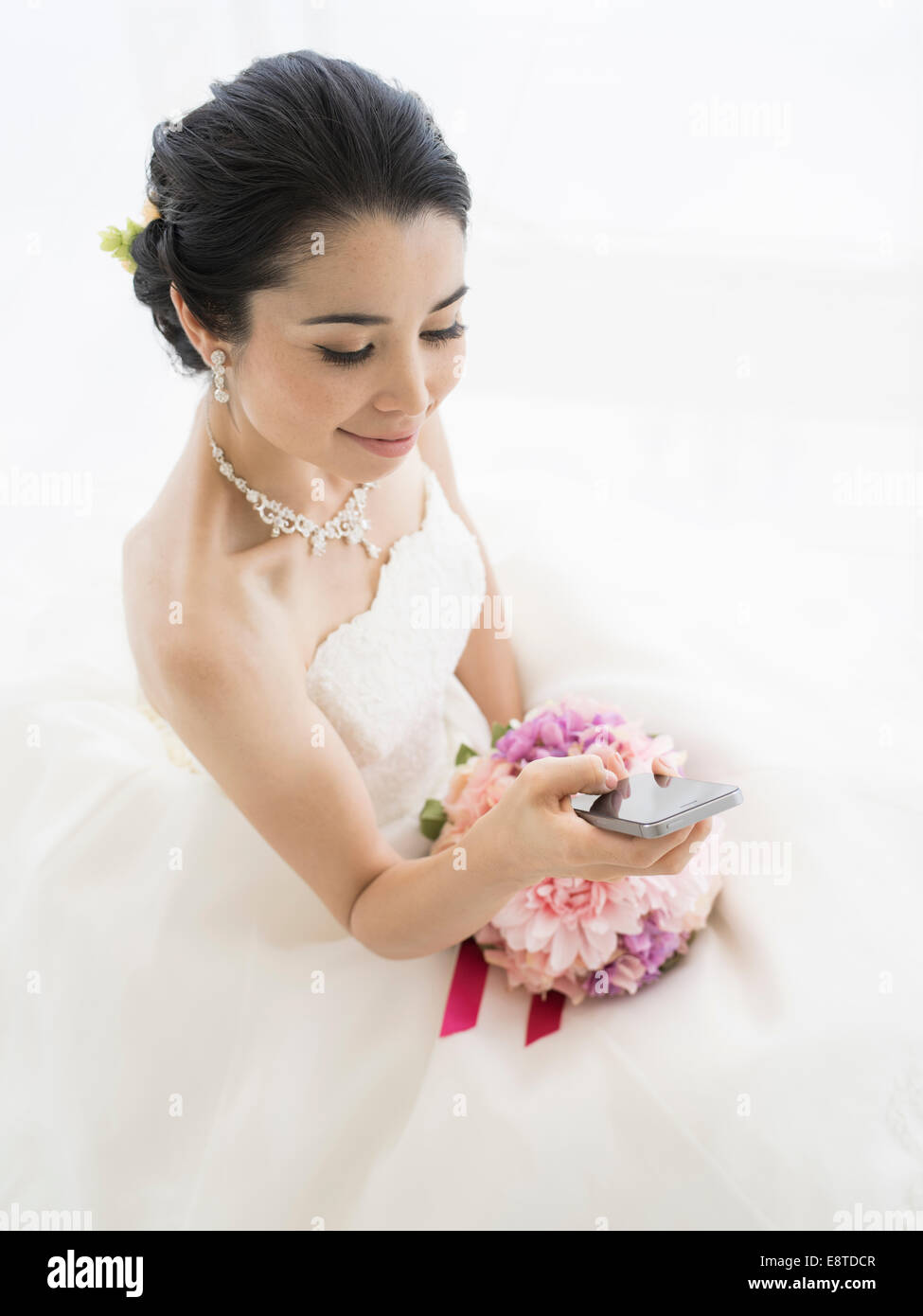 Mixed race, Asian / American bride in white wedding dress using iphone to text friends Stock Photo