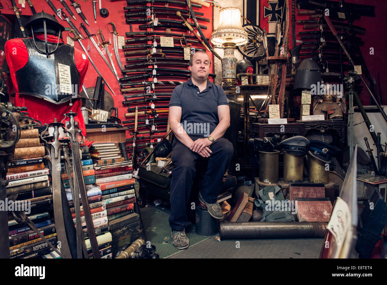 A male shopkeeper stands in his historical military antique shop surrounded by guns, swords, rifles, armor etc. Stock Photo