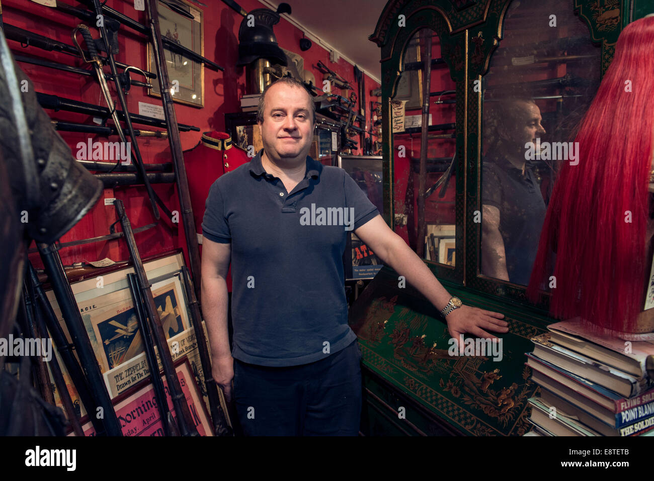 A male shopkeeper stands in his historical military antique shop surrounded by guns, swords, rifles, armor etc. Stock Photo