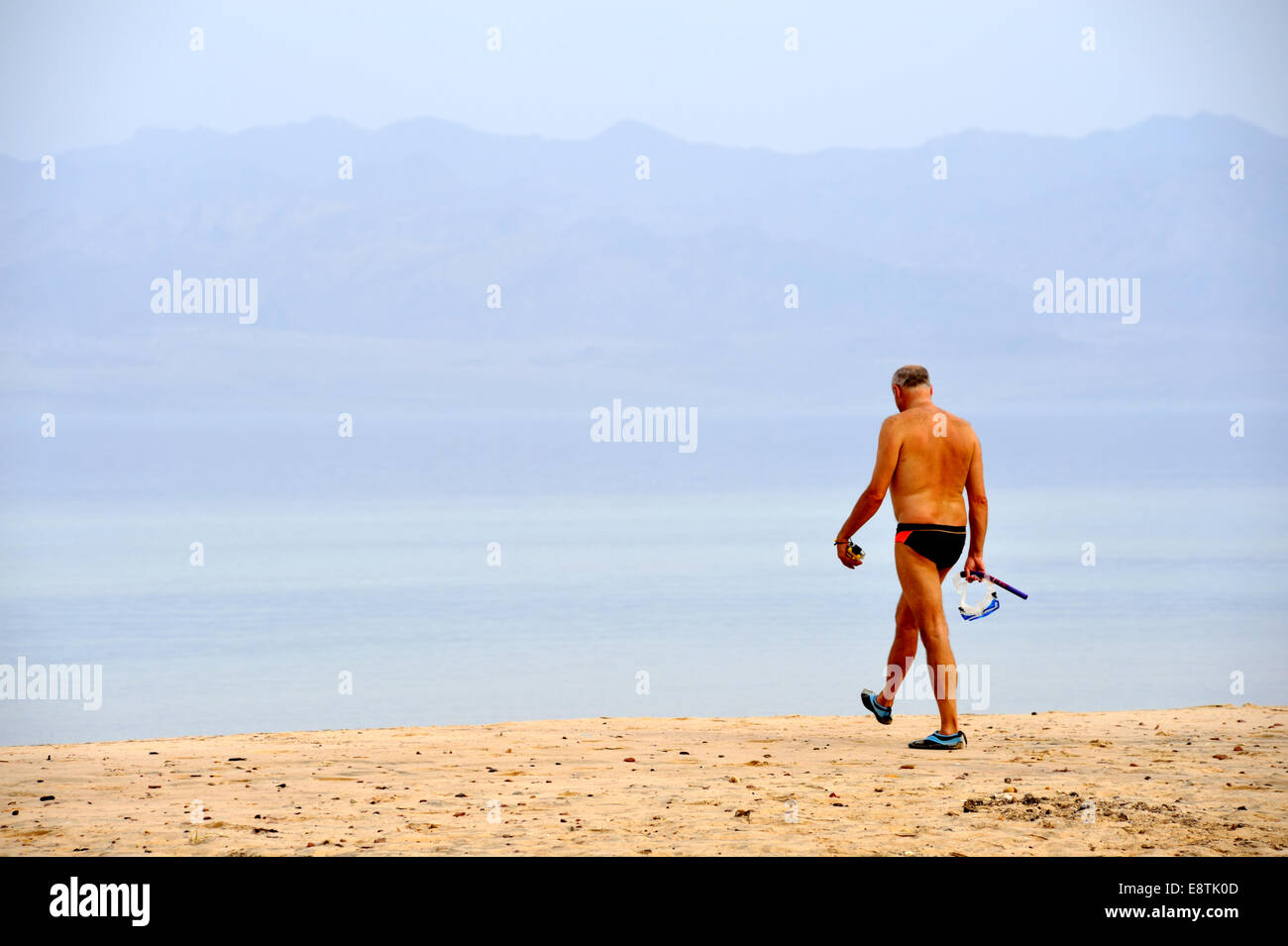 Man in swim suit carrying mask and snorkel walking across sandy beach to sea Stock Photo