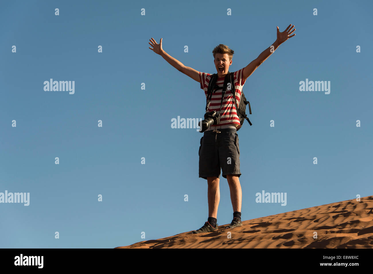 Teenager with photo camera standing on a dune and spreading his arms, Sossusvlei, Namib Desert, Namibia Stock Photo