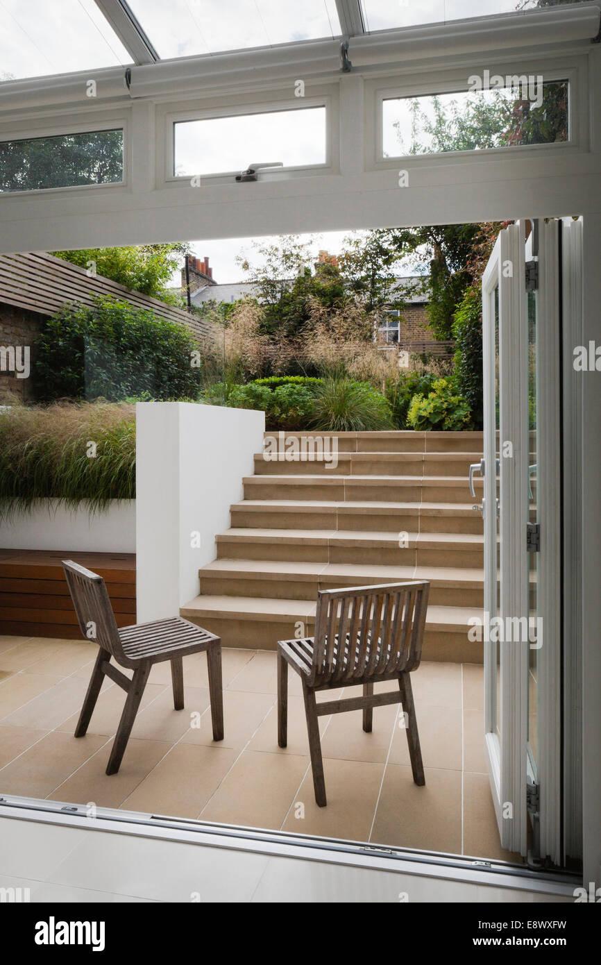 French windows open onto the lower York stone paved outside area, with benchstore and York stone steps leading up to the larger, upper level. Contemporary, teak chairs designed by Matthew Hilton for Habitat. Rear garden in Greenwich, South East London, UK Stock Photo