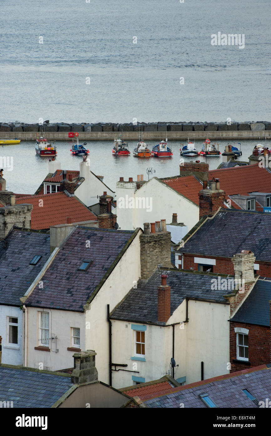 High view over rooftops of tiny, quaint cottages, to boats in the harbour & sea beyond - seaside village of Staithes, North Yorkshire, England, UK. Stock Photo