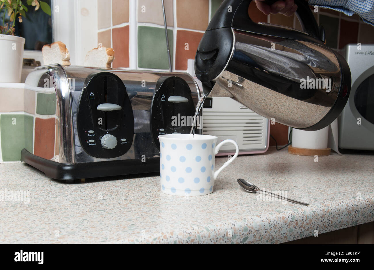 Kettle pouring boiling water into a tea cup in a kitchen with a toaster in the  background Stock Photo