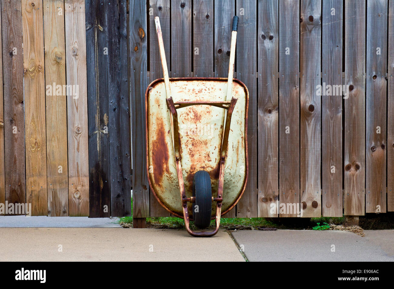 Old rusty wheelbarrow leaning vertically on a wooden privacy fence. Stock Photo