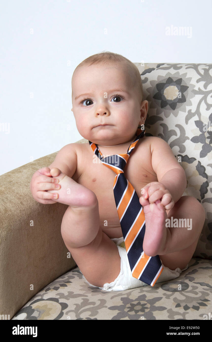 9 month old baby boy sitting on a chair, holding his toes, wearing a tie and a diaper Stock Photo