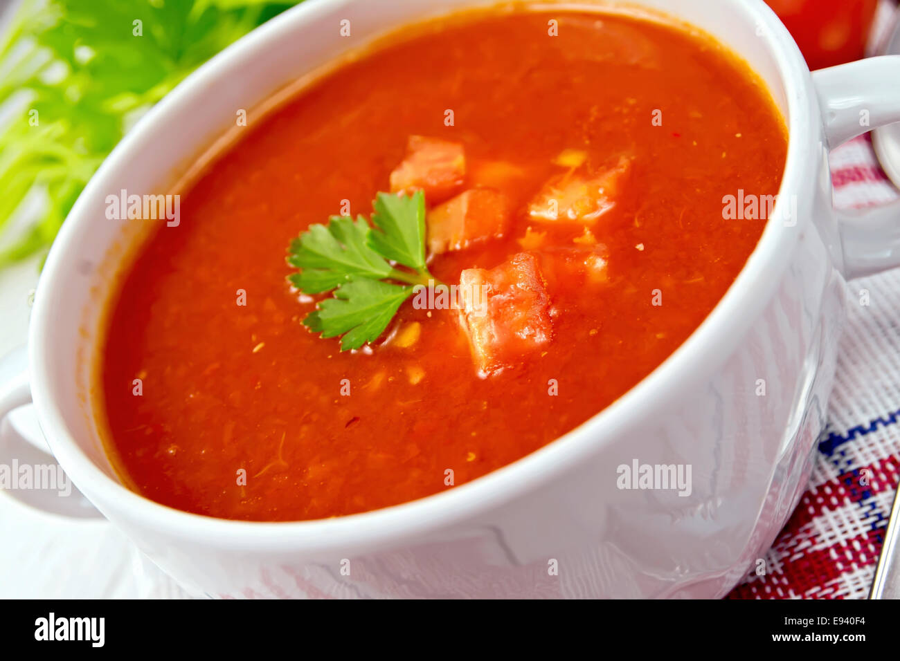 Tomato soup with chunks of vegetables in a white bowl on a napkin with a spoon, tomatoes, parsley on a lighter background board Stock Photo