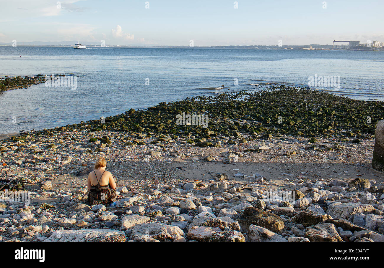 Young woman sitting on the stone beach writing during evening at Praça do Comércio,  Baixa Pombalina, Lisbon, Portugal Stock Photo