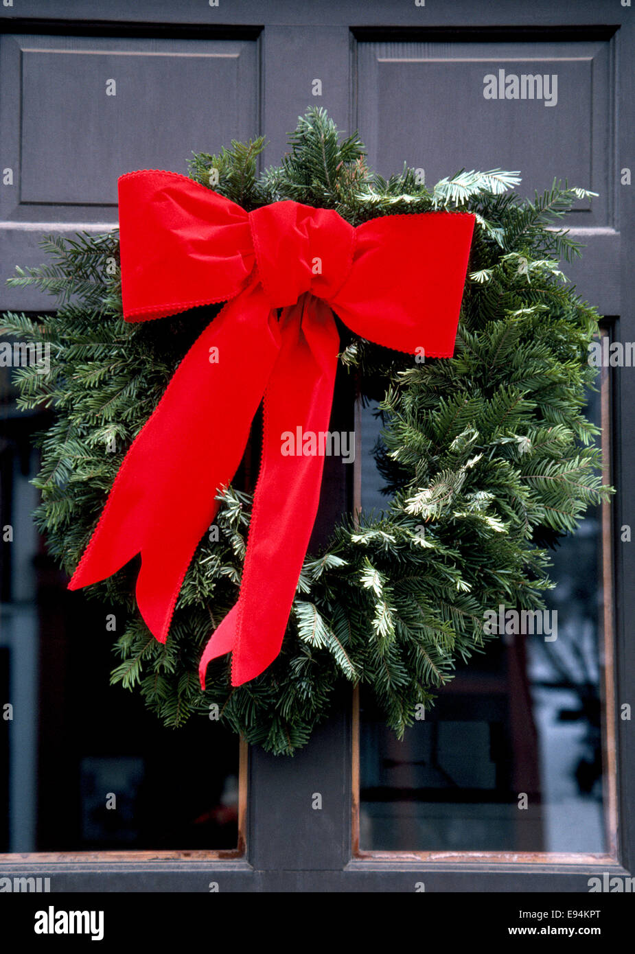 A big bright red holiday bow attached to a wreath of evergreens decorates the entrance door to a house in the USA at Christmastime. Stock Photo