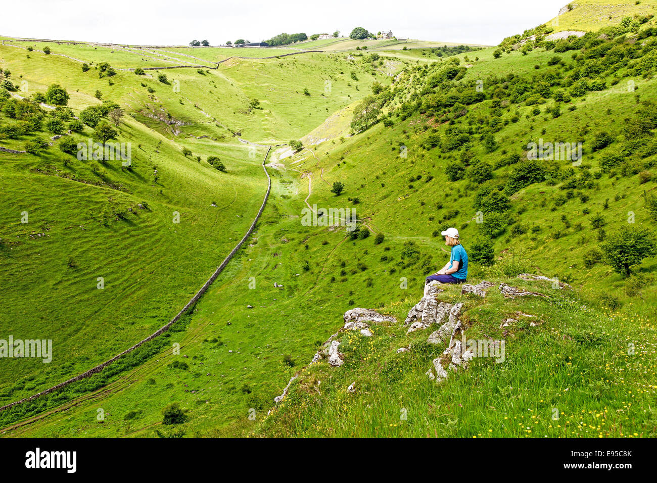 A woman sitting dangling her legs on a rock overlooking Cressbrook Dale Derbyshire Peak District National Park England UK Stock Photo