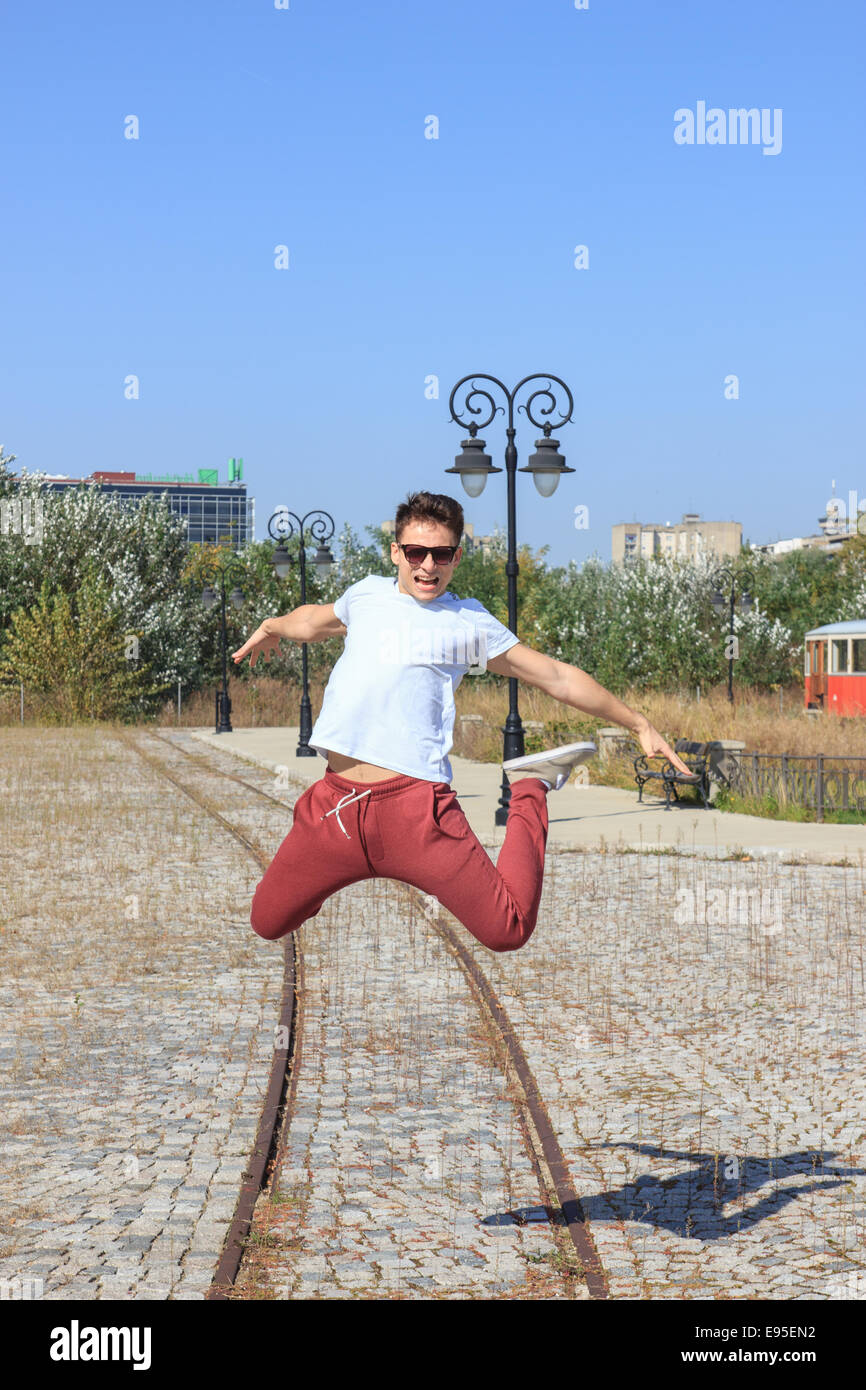 smiling happy male teenager jumps in retro park and wearing sunglasses in red sweatpants and white t-shirt Stock Photo