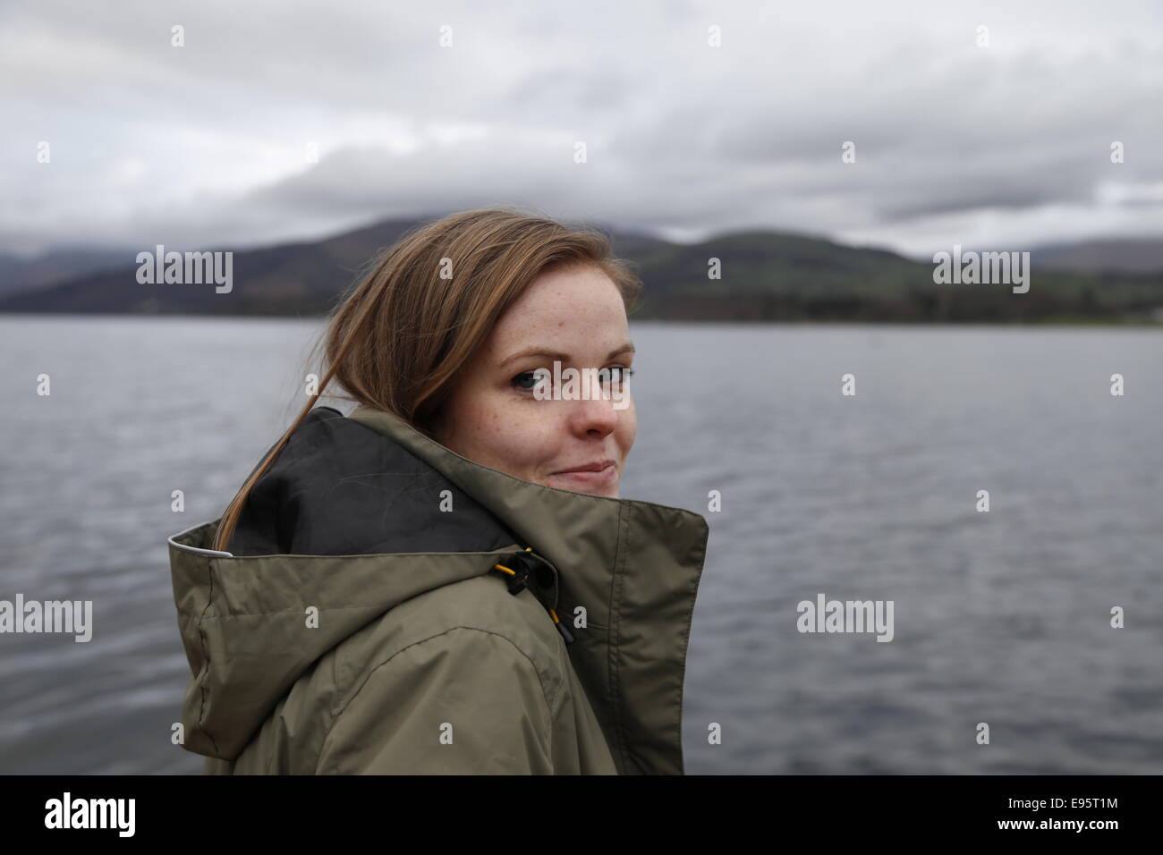 Pretty young woman wearing a coat stands smiling at the shores of Lake Windermere. Stock Photo