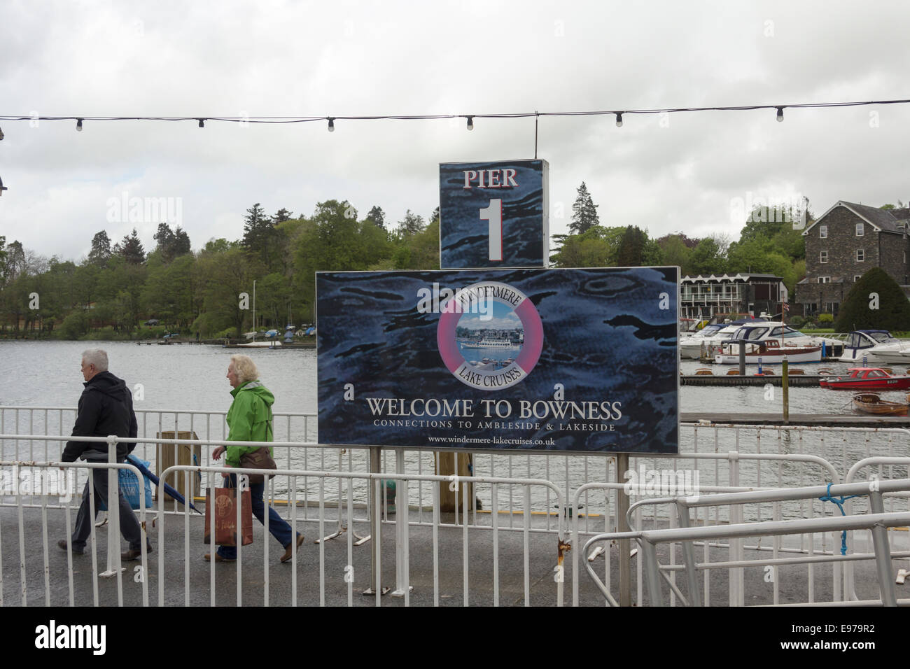 Two adult people at 'Welcome to Bowness' sign on the Windermere Steamer pier at Bowness-on-Windermere on a dull and rainy day. Stock Photo