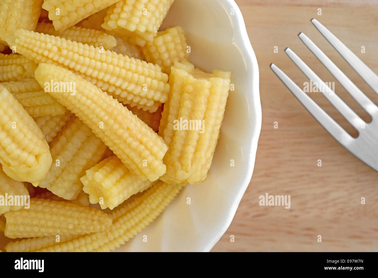 A very close view of small baby corn nuggets in a small bowl with a fork to the side on a wood table top. Stock Photo