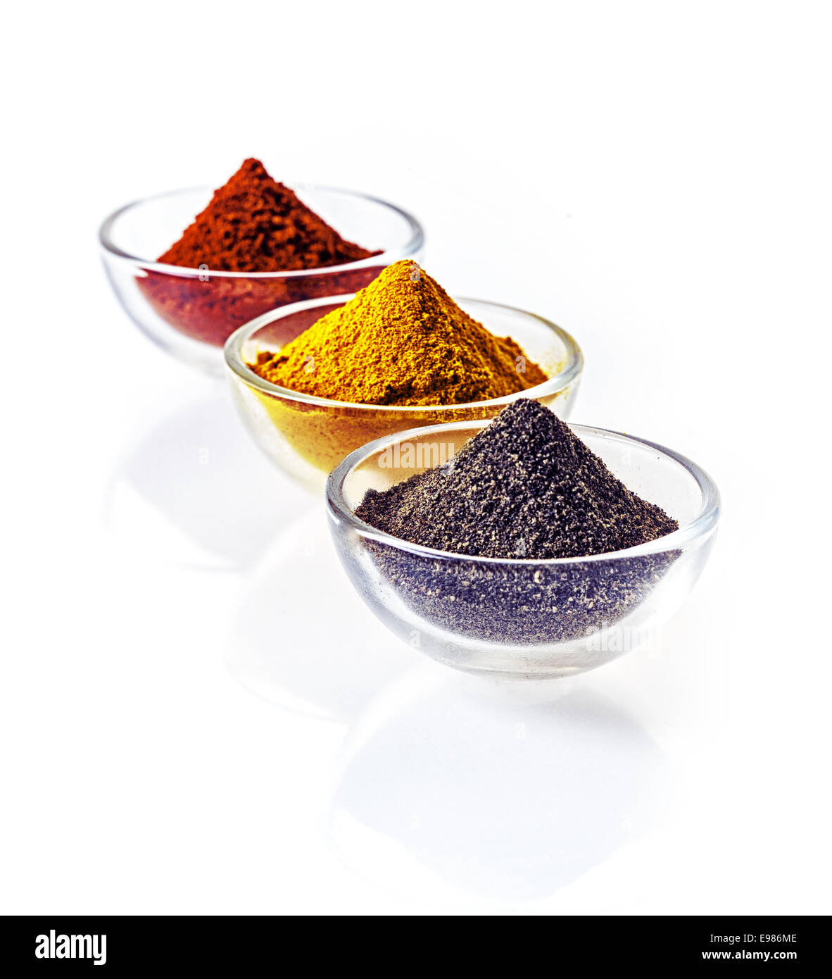 Three clear glass bowls of colourful of ground spice and black lava salt in conical heaps arranged in a receding row on a white Stock Photo
